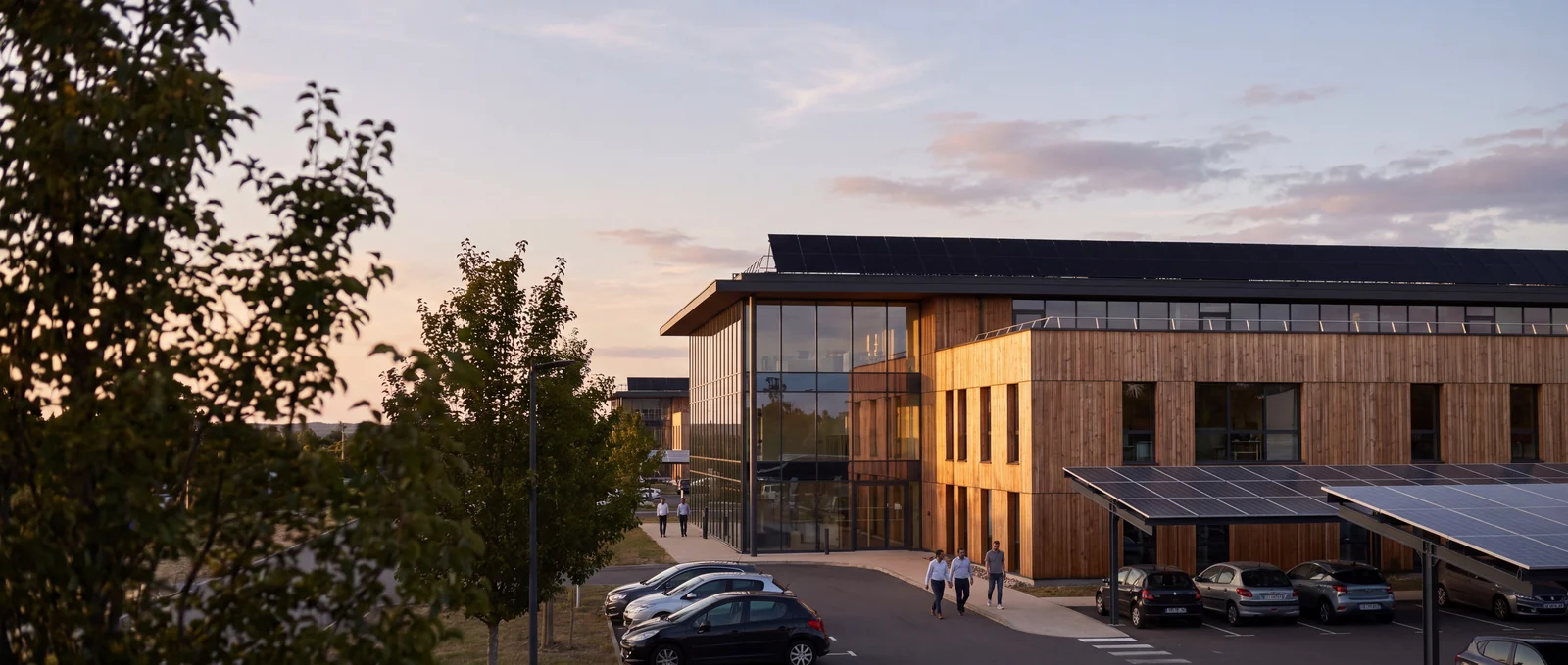 Illustration : immeuble de bureaux rénové en fin de journée : façade bois et verre, panneaux solaires en toiture et ombrières photovoltaïques sur le parking